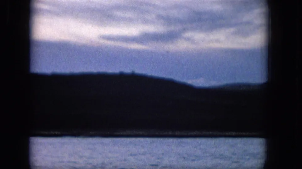 1965 8mm frame looking through a dark window frame toward rippled water, a silhouetted hillside or shoreline, and a soft blue sky with streaked clouds.