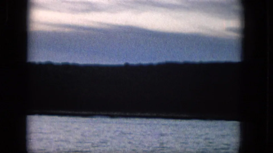 1965 8mm frame showing a dark foreground window frame around a calm body of water, a low silhouetted shoreline, and pale clouds over blue sky.