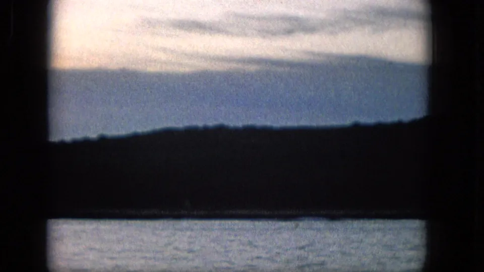 1965 Super 8 frame from supplied Molokai metadata showing calm water, silhouetted land, and a pale blue sky divided by dark frame bars.
