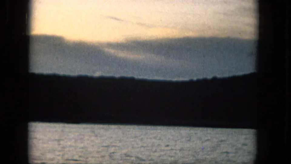 1965 Super 8 frame from supplied Molokai metadata with a warm sky, distant layered land, and calm water seen through dark framing.