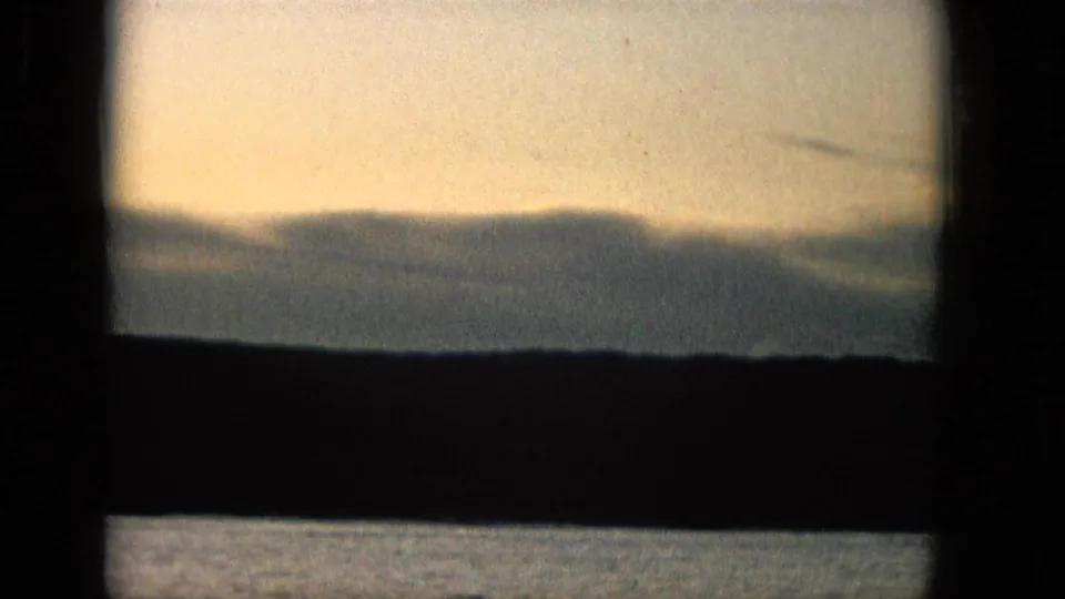 1965 Super 8 frame from supplied Molokai metadata showing a muted yellow sky, silhouetted shoreline, and water beneath dark window-like bands.