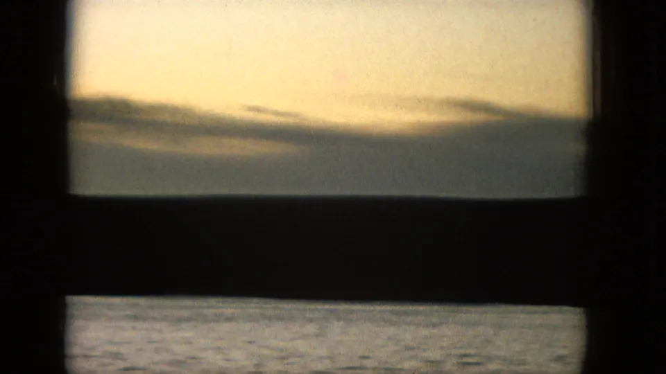 1965 United States 16mm home-movie frame showing a calm water horizon and glowing sunset sky viewed through a dark window or railing structure.