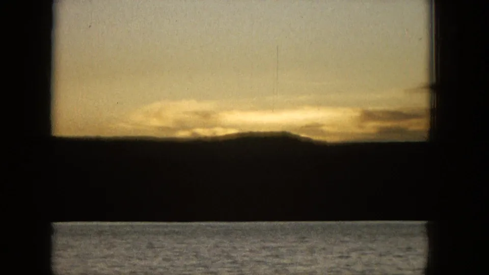 1965 United States 16mm home-movie frame showing a low shoreline silhouette, golden sunset light, and a dark window or railing edge in the foreground.