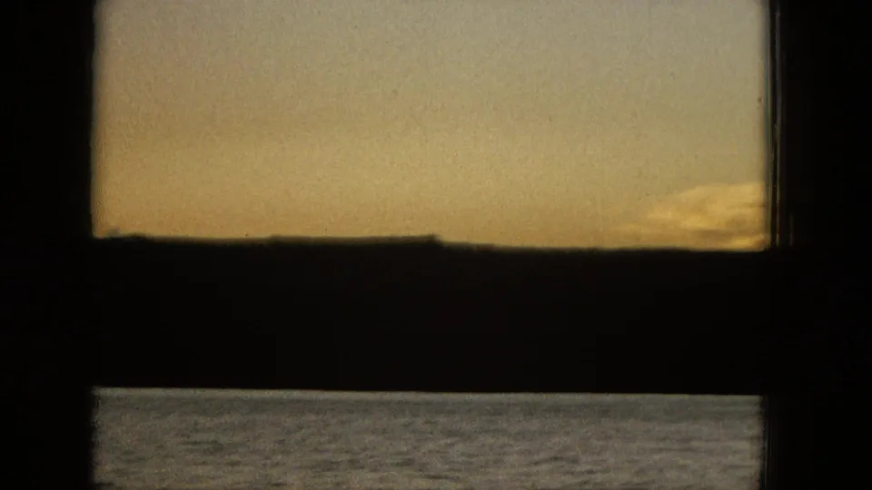 1965 United States 16mm home-movie frame with pale evening sky, a low shoreline silhouette, and dark foreground framing bars.