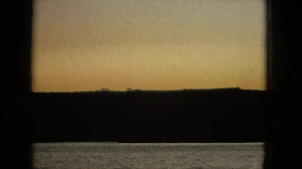 A calm body of water and dark low shoreline are seen through a heavy window frame under a warm orange sky, supported by metadata as 1965 archival footage.