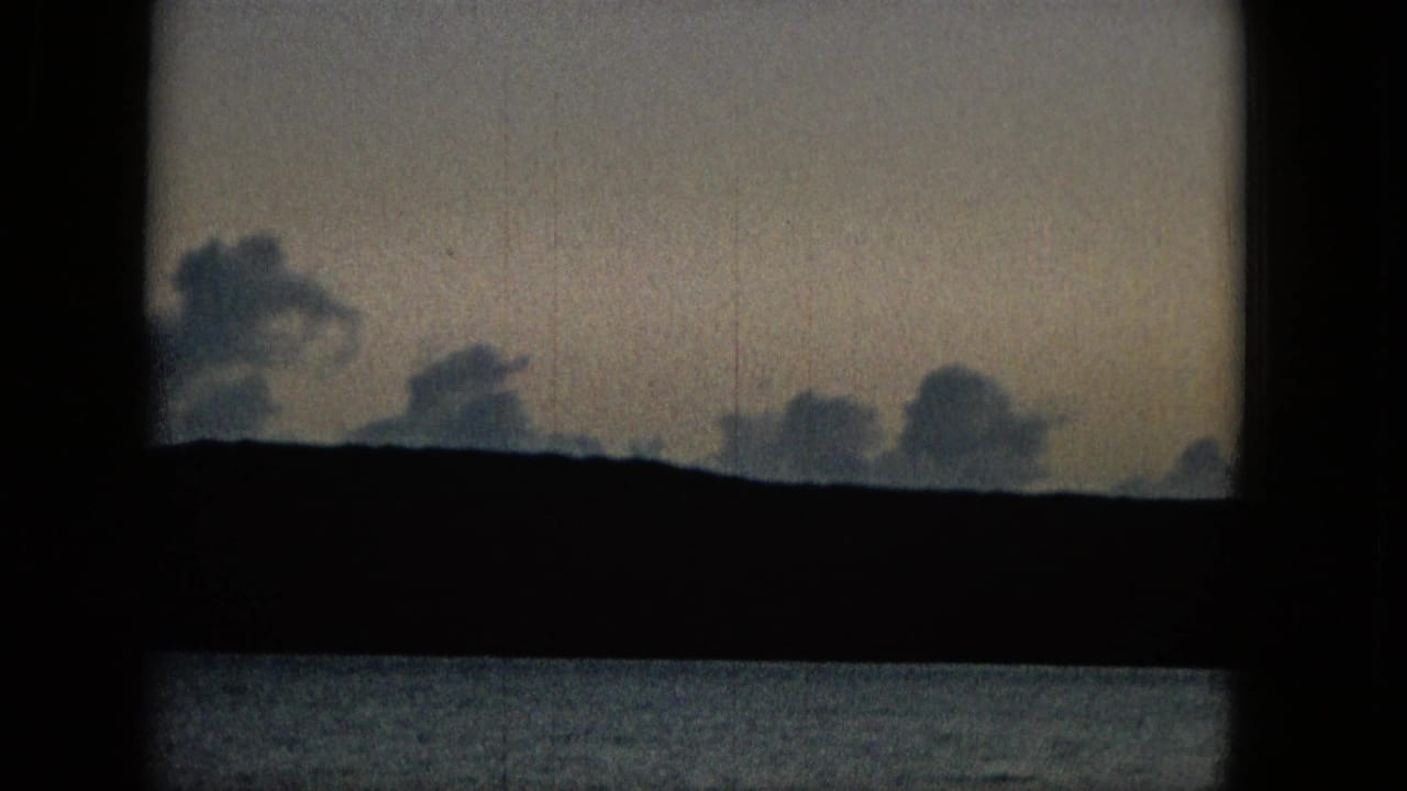 Soft clouds sit above a dark landmass and calm water, framed by the black crossbars of a window in vintage home-movie footage.