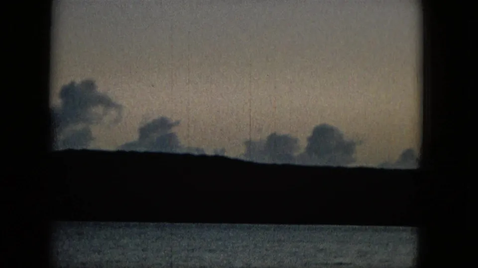 Soft clouds sit above a dark landmass and calm water, framed by the black crossbars of a window in vintage home-movie footage.