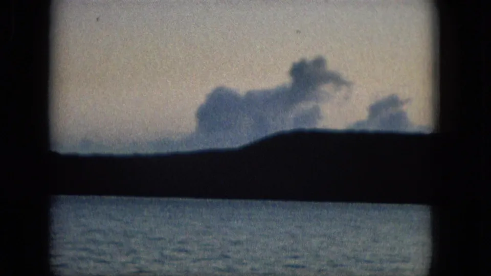 A blue-gray coastal horizon with low clouds and water appears through the window frame, with visible grain and muted color from the archival film scan.