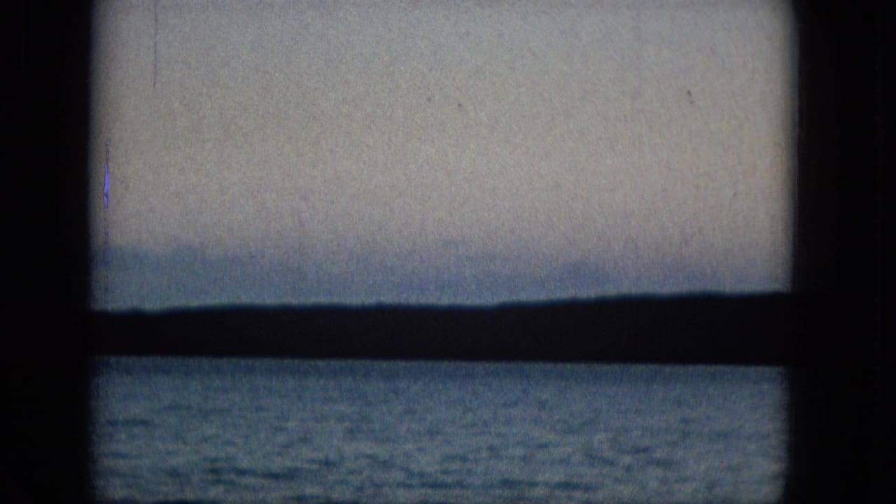 A 1960s archival home-movie frame shows a seascape viewed through a dark window frame, with blue water, distant landforms, and a pale hazy sky.