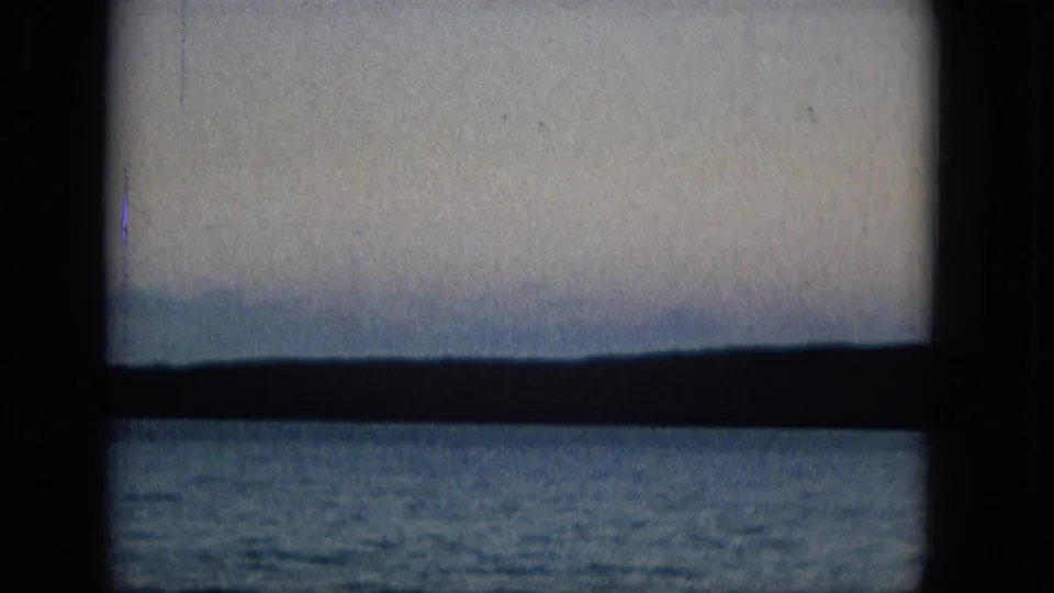 A 1960s archival home-movie frame shows a seascape viewed through a dark window frame, with blue water, distant landforms, and a pale hazy sky.