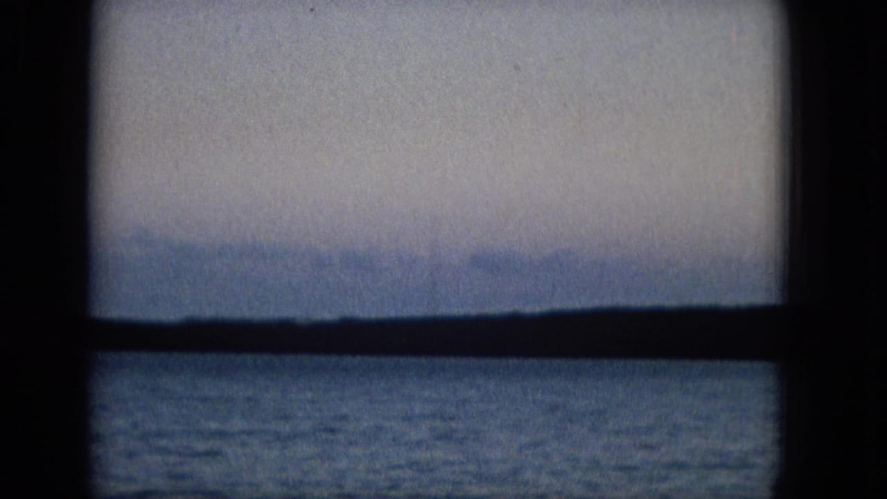 A dark window grid frames open water and a distant hazy mountain or shoreline, consistent with archival island travel footage from the 1960s metadata.