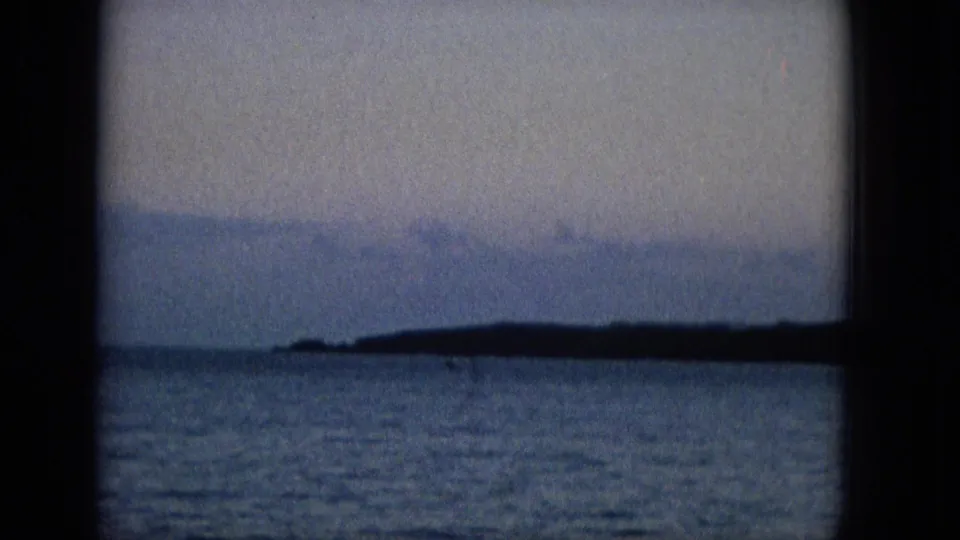 The frame shows blue water, a low dark shoreline, and pale sky viewed through the lower panes of a window in soft, grainy archival footage.