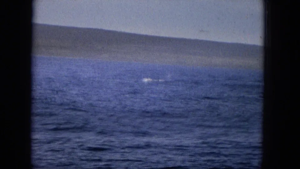 Archival home-movie view across blue water toward a low coastline, with a small splash or disturbance visible near the surface.