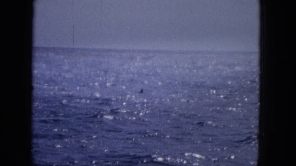 1965 archival home-movie frame showing blue ocean water, a pale horizon, and dark vertical window framing with no people visible.