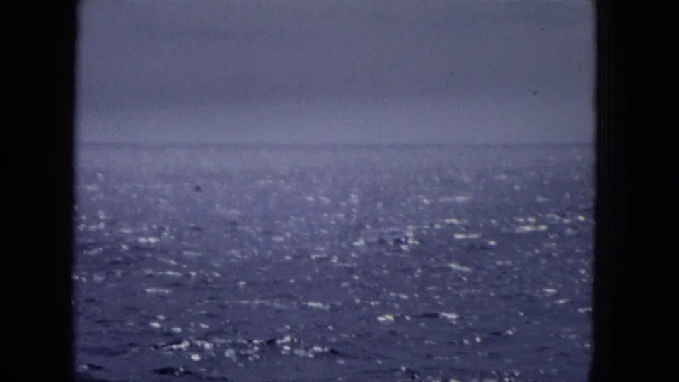 1965 archival home-movie frame showing a calm blue ocean horizon, pale sky, and visible film texture through a dark window frame.