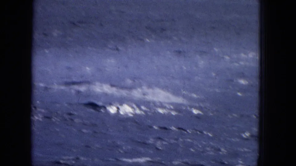 Soft, blue-tinted 8mm frame showing a whale or large marine animal moving through the ocean and leaving a stretched white splash pattern on the surface.