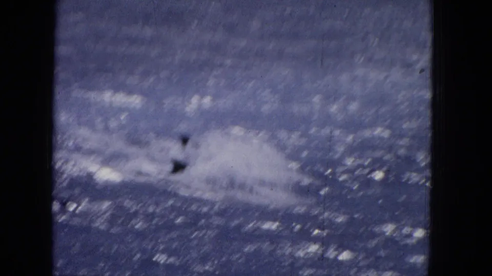 Final 1965 8mm frame with a larger blurred splash and dark marine shape at the water surface, retaining visible film grain and a strong blue color cast.