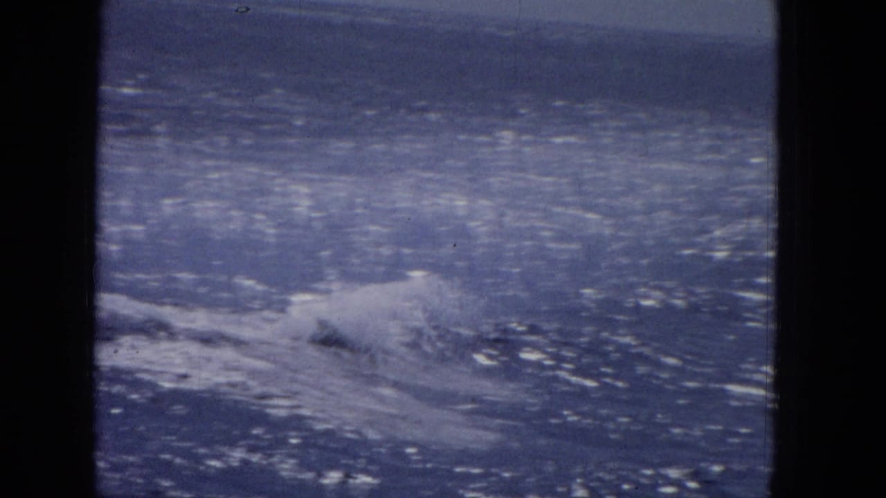 1965 archival home-movie frame showing blue ocean water and wake from a boat viewpoint, with heavy dark gate edges and a soft horizon.