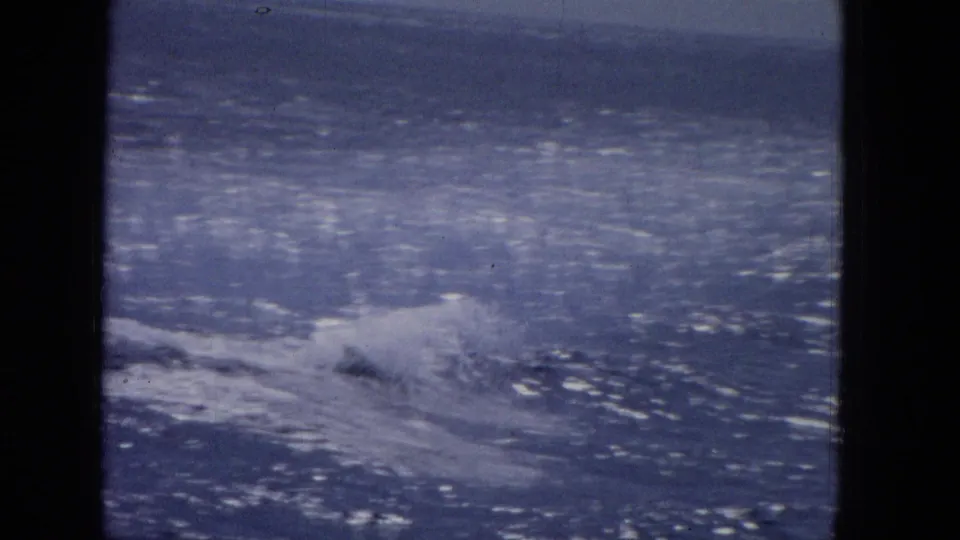 1965 archival home-movie frame showing blue ocean water and wake from a boat viewpoint, with heavy dark gate edges and a soft horizon.
