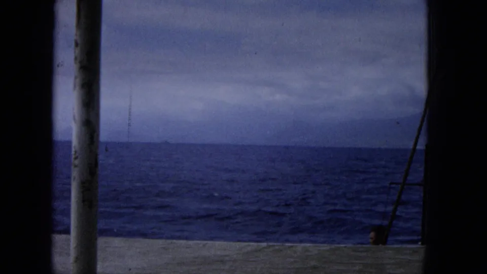 Archival home-movie view from a boat deck toward open blue water and an overcast horizon, with railing and a vertical post framing the scene.
