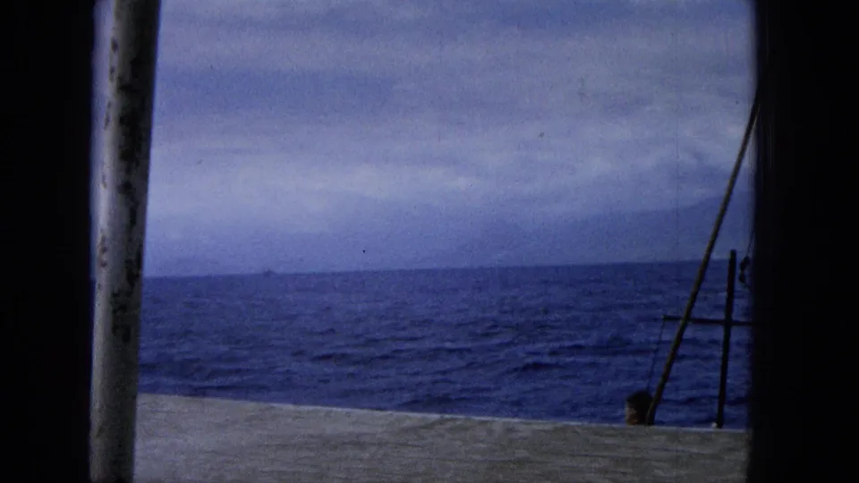 1965 home-movie frame looking across a boat deck toward the ocean and low horizon, with a post, rigging, and a small person partly visible near the lower right.