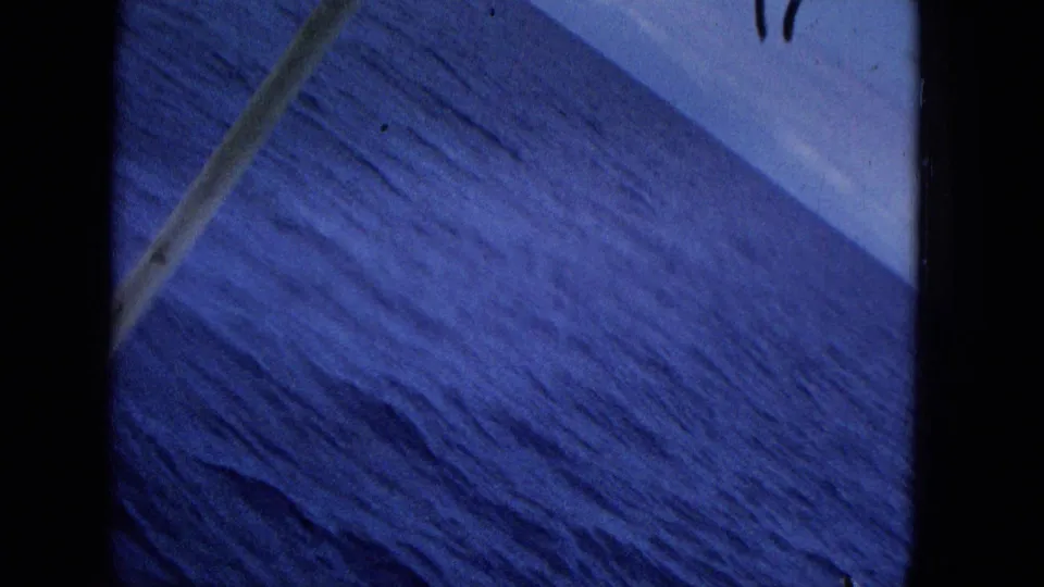 Tilted archival boat-view frame of textured blue ocean water, crossed by a diagonal structural element and bordered by dark film edges.