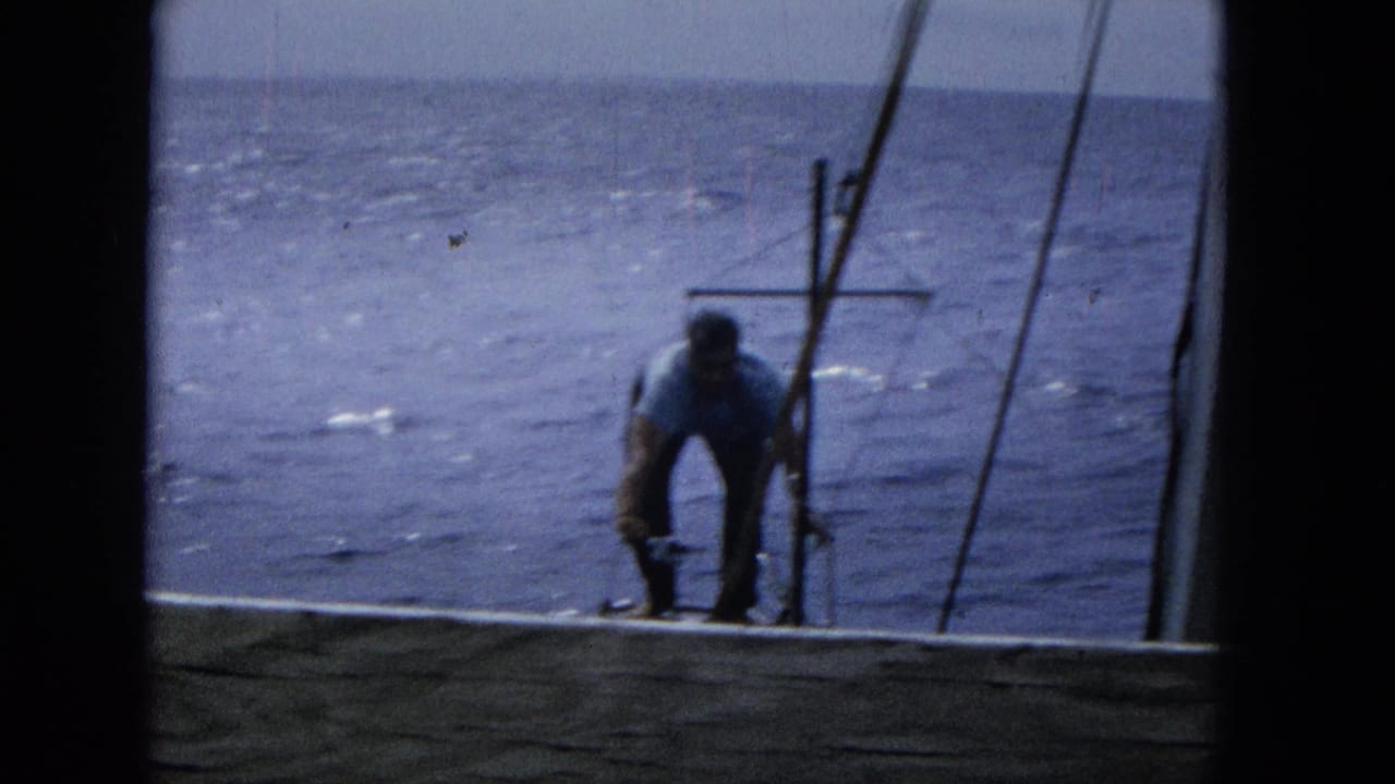 1965 Super 8 frame showing a man in a light shirt bending near sailboat rigging, seen through a dark window or hatch frame with open ocean behind him.