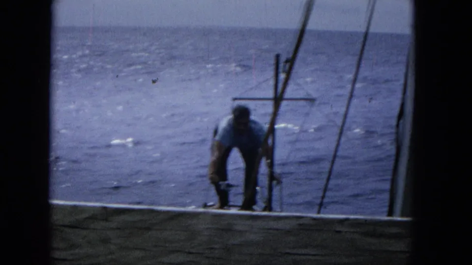 1965 Super 8 frame showing a man in a light shirt bending near sailboat rigging, seen through a dark window or hatch frame with open ocean behind him.