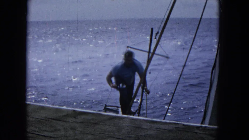 1965 Super 8 frame of a man standing on or beside the sailboat deck near rigging, viewed through a dark frame with sunlit ocean behind him.