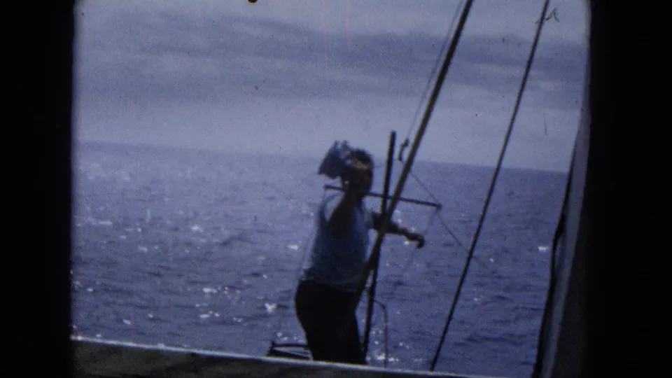 1965 Super 8 frame showing a man on a sailboat holding a small camera or optical device near his face, with rigging and open ocean visible behind him.