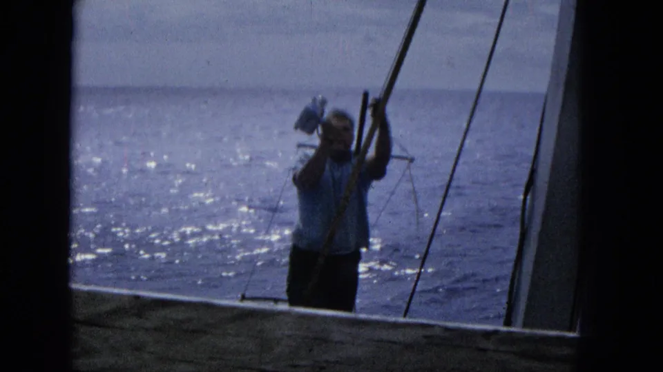 1965 Super 8 frame of a man on a sailboat deck lifting one arm near the rigging, with sparkling ocean and a low horizon in the background.
