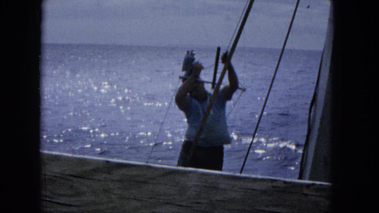 A man in casual clothing stands near boat rigging with a camera, framed by dark structural edges against sparkling Pacific water in 1965 Super 8 footage.