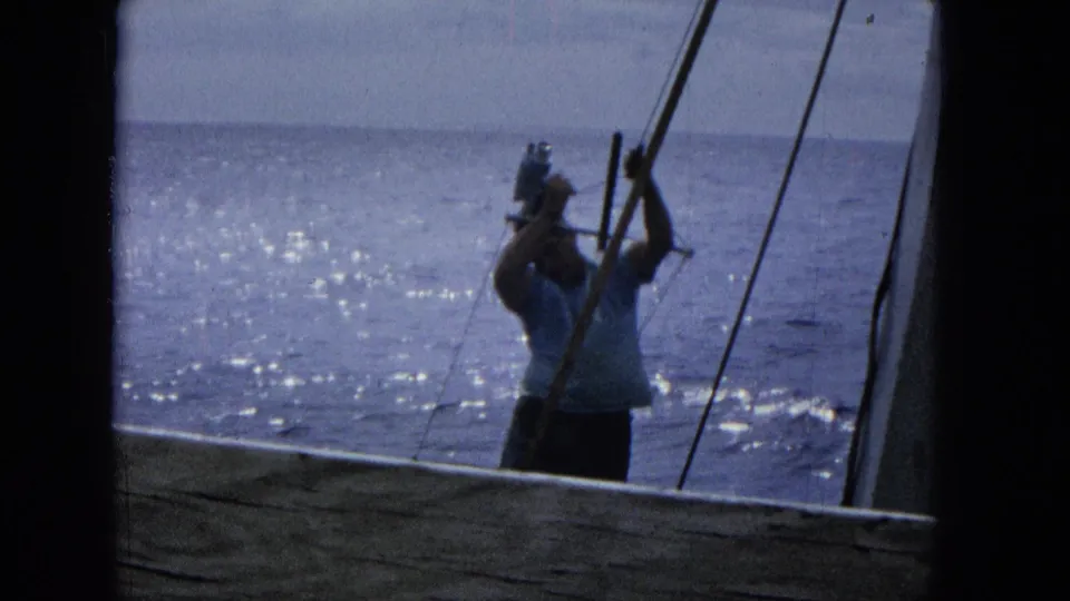 A man in casual clothing stands near boat rigging with a camera, framed by dark structural edges against sparkling Pacific water in 1965 Super 8 footage.