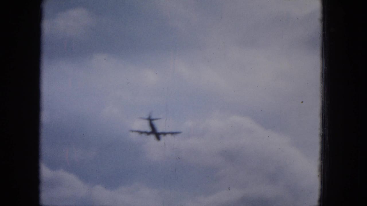A distant military-style aircraft appears in soft focus against a pale cloudy sky in the 1965 home-movie footage.