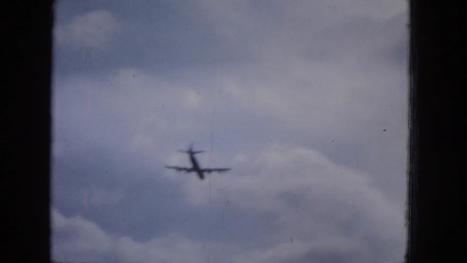 A distant military-style aircraft appears in soft focus against a pale cloudy sky in the 1965 home-movie footage.