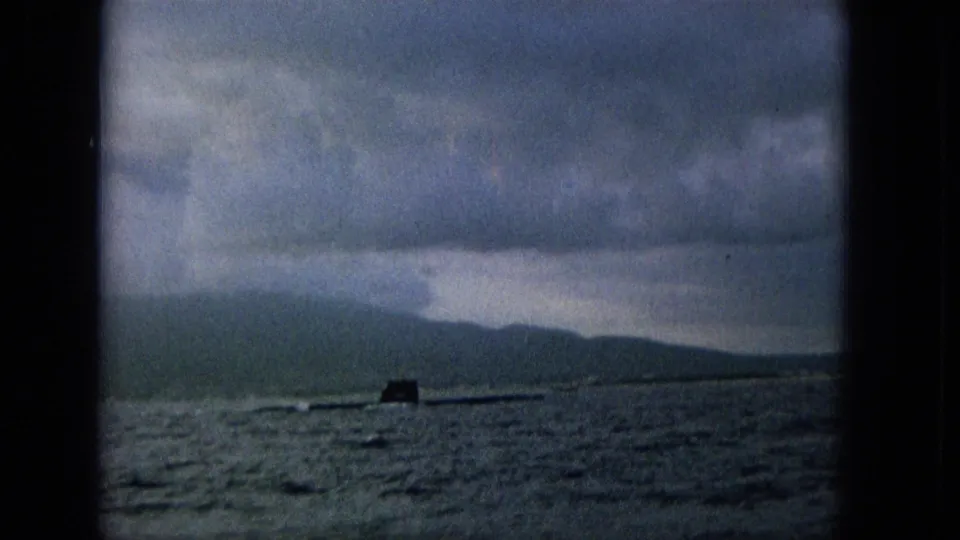 A partially submerged submarine appears on choppy water with a hazy island mountain coastline in the background.