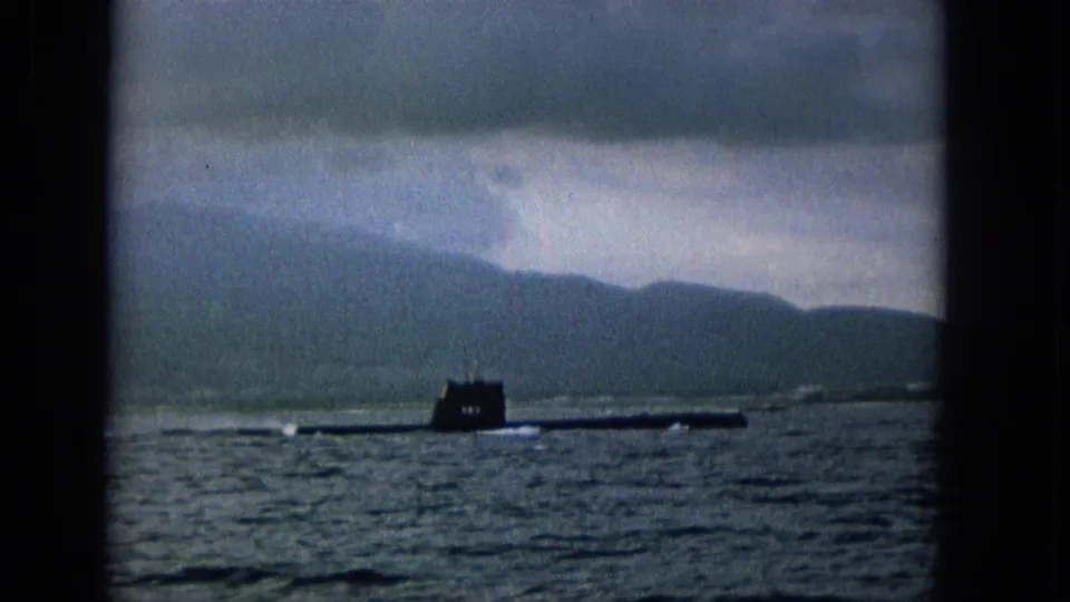 The submarine is seen more clearly across the water, framed by a distant island coastline and overcast sky in 1965 8mm footage.