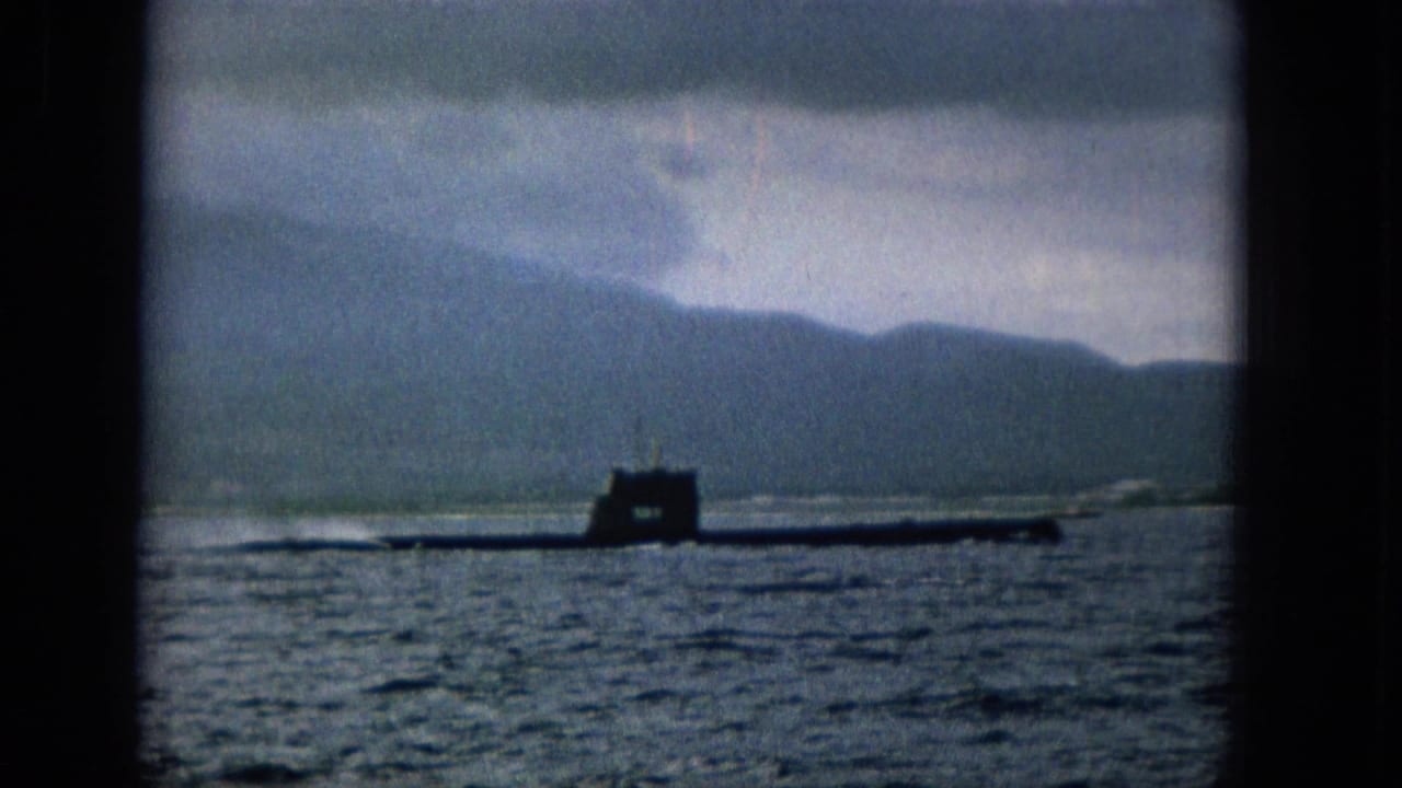 1965 home-movie frame showing a dark submarine moving across water in Hawaii metadata context, with distant mountains, cloudy sky, and heavy dark frame edges.