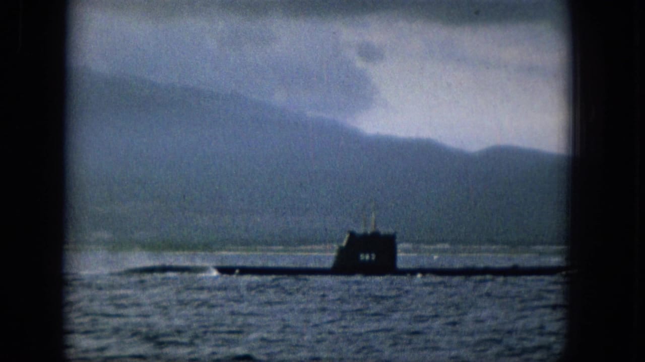 The submarine appears closer to the right side of the frame, with its conning tower and long hull visible on the water below a hazy mountain ridge.