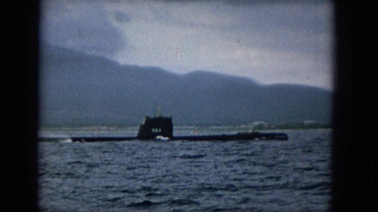 1965 home-movie frame of a dark submarine moving across Molokai waters, seen through a dark window-like border with island hills in the distance.