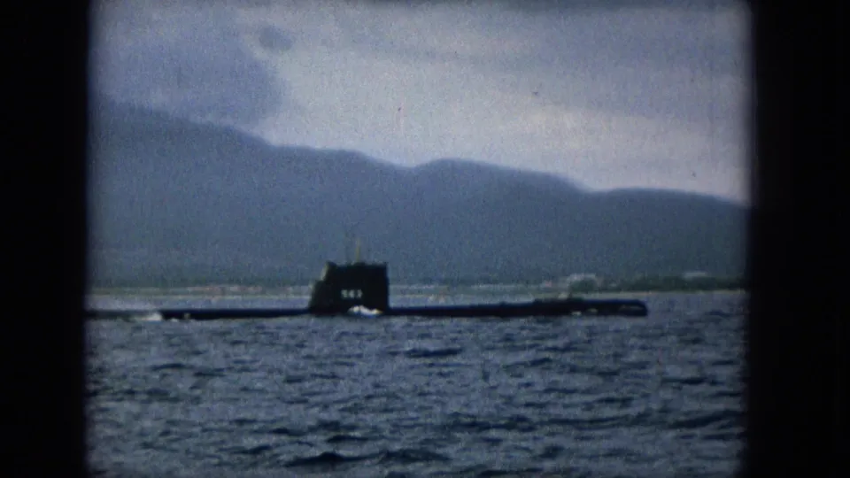 1965 home-movie frame of a dark submarine moving across Molokai waters, seen through a dark window-like border with island hills in the distance.