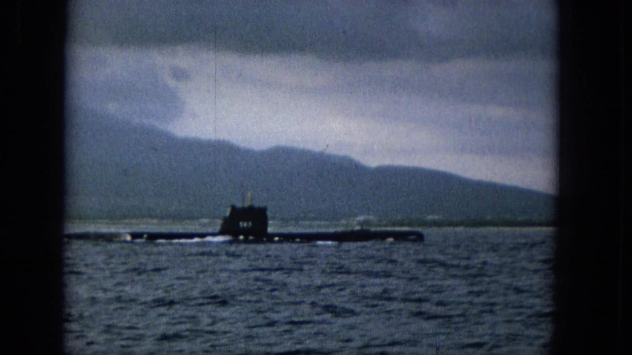 A dark submarine silhouette cruises across calm Hawaiian waters, with Molokai hills and a gray cloud layer visible beyond the shoreline.