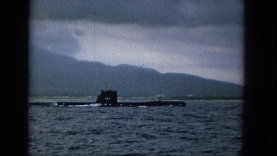 A dark submarine silhouette cruises across calm Hawaiian waters, with Molokai hills and a gray cloud layer visible beyond the shoreline.