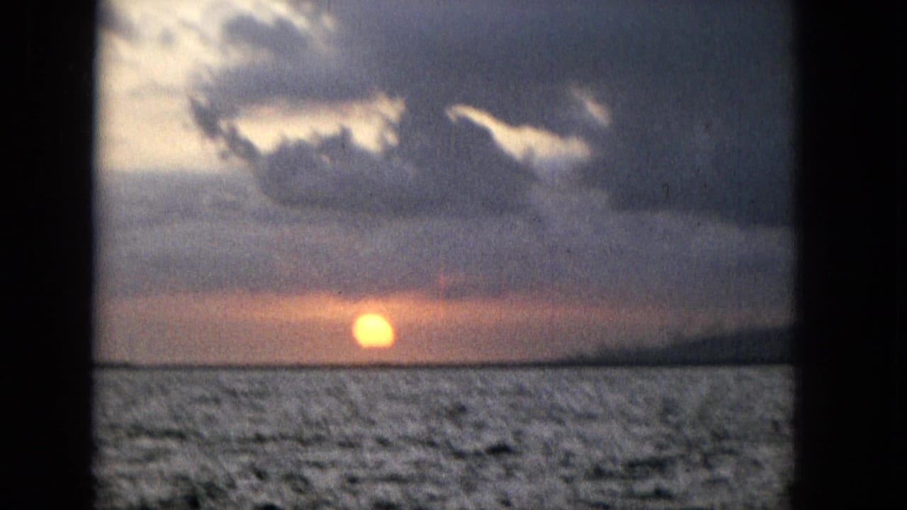 1965 Super 8 frame showing a golden sunset over rippled ocean water, with heavy clouds and dark window-frame bars crossing the view.