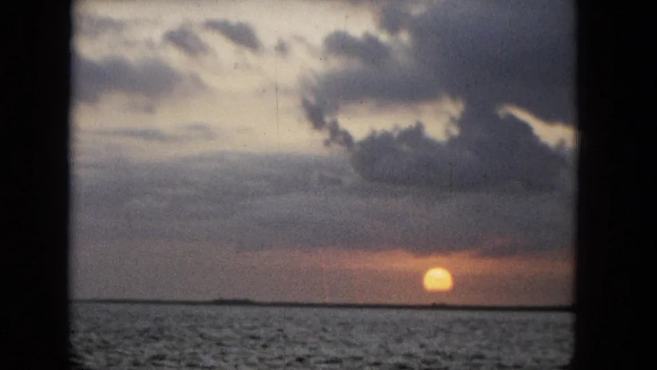 1965 Super 8 frame with a warm sun near the ocean horizon, layered storm clouds overhead, and dark window-frame edges around the scene.