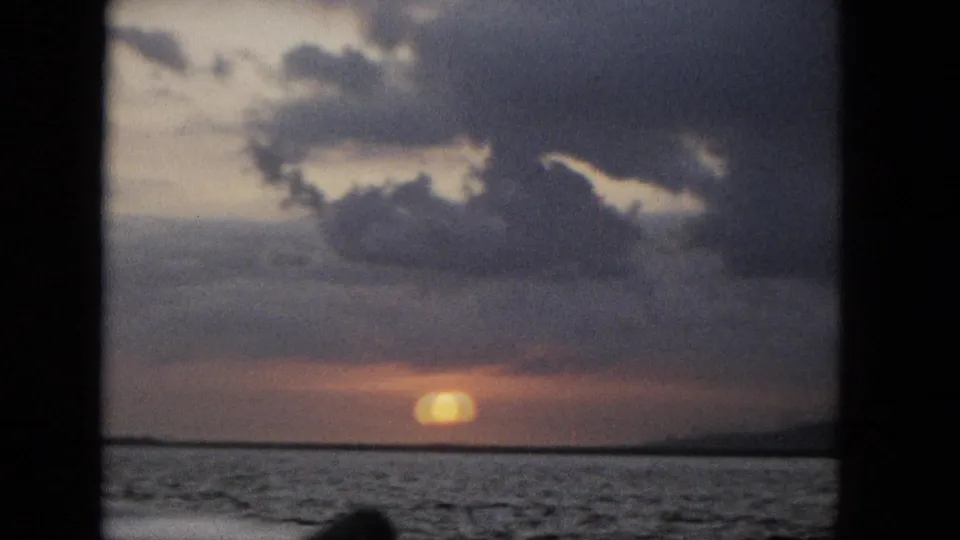 1965 Super 8 sunset frame showing a softened golden sun at the horizon, dark rippled water, brooding clouds, and black window-frame borders.