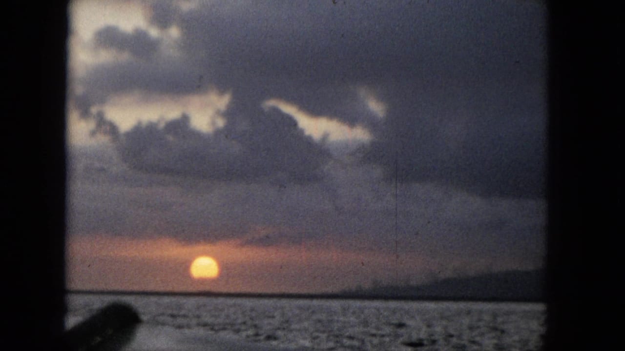 1965 home-movie frame showing an orange sun low over water under heavy clouds, with dark vertical framing at the edge of the view.