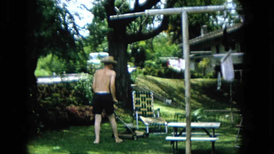 1965 Super 8 frame of a shirtless man wearing a light hat and pushing a mower through a green backyard with outdoor furniture and a clothesline nearby.
