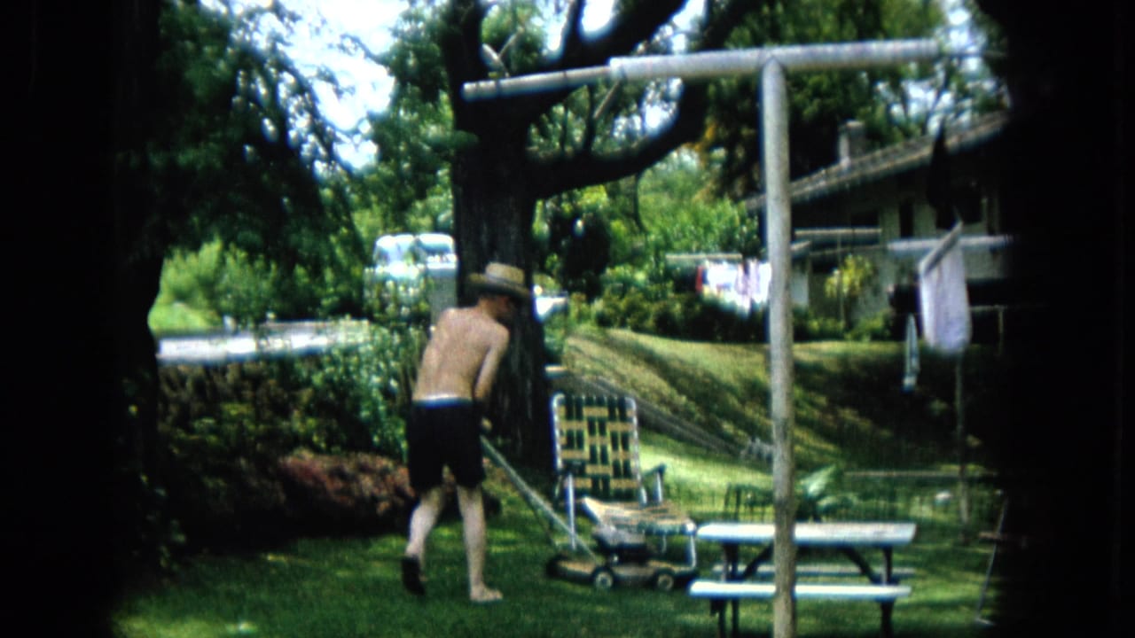 1965 U.S. home-movie frame showing a shirtless man in a straw hat mowing grass near a picnic table, lawn chair, clothesline, shrubs, and a large tree.