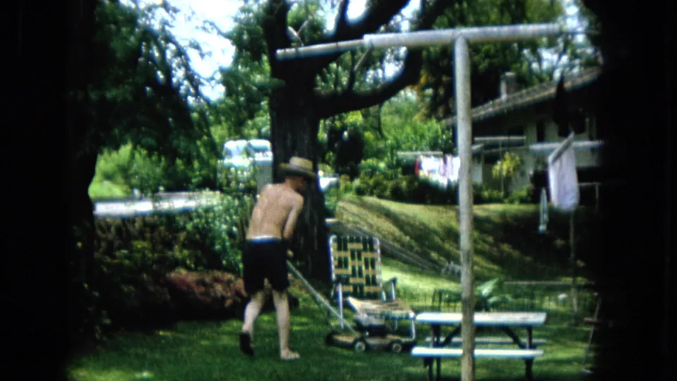1965 U.S. home-movie frame showing a shirtless man in a straw hat mowing grass near a picnic table, lawn chair, clothesline, shrubs, and a large tree.